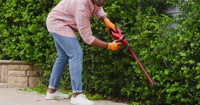 African American man walking along paved walkway using red trimmer in orange gloves removing growth