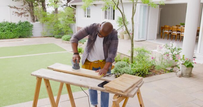 After clamping, African American man wearing toolbelt sanding plank on patio with sander to finish