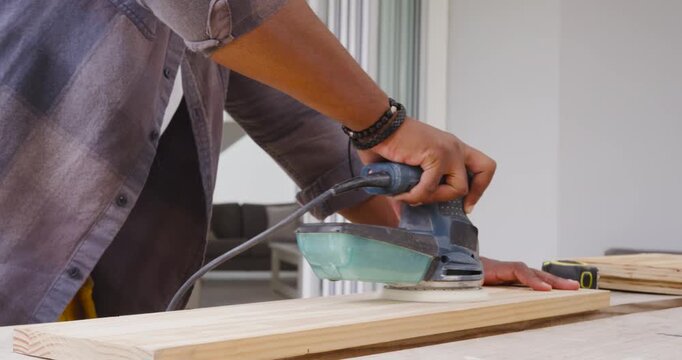 African American man powering corded sander guiding plank on home bench with goggles for finishing