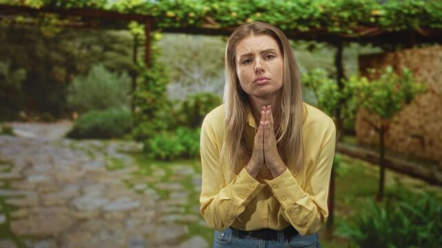 Woman with hands clasped in prayer and puckered lips pleading in a sunlit stone courtyard, wearing yellow shirt and jeans; pleading apology.