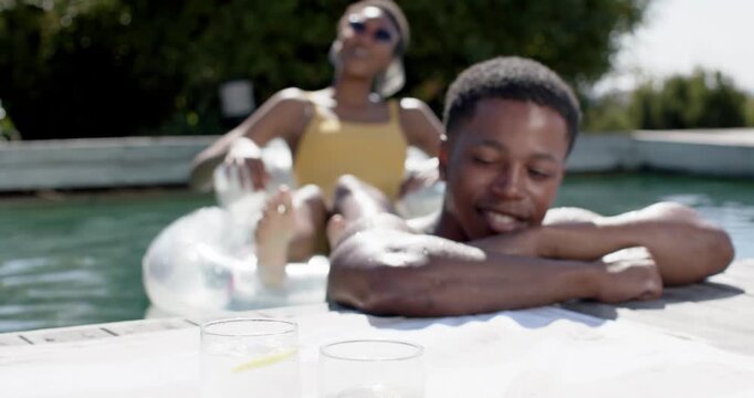 Couple at pool edge, man leaning smiling, woman reclining in yellow swimsuit soaking sun, glasses