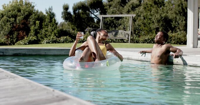 African American couple smiling, woman in yellow swimsuit raising drink in clear ring at home pool