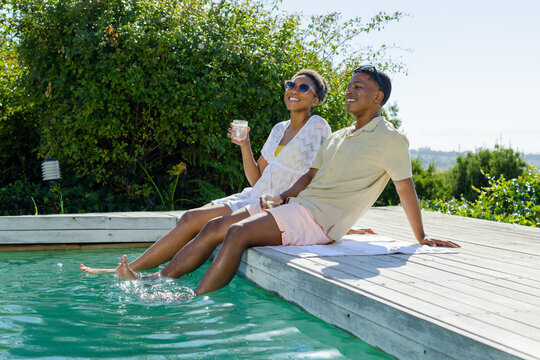 African American couple sitting on white towel on wooden deck, dipping feet and holding drinks