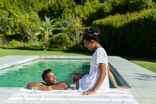 African American couple lounging by pool on deck, both wearing swimwear, striped towel, two glasses