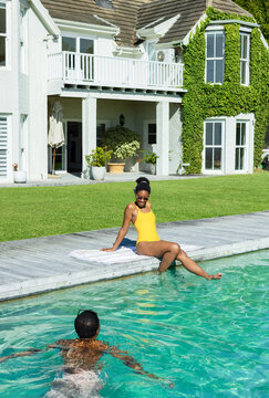 Diverse friends sitting on deck by pool, wearing yellow swimsuit and sunglasses, other swimming