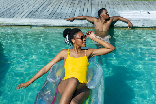 African couple lounging at pool woman reclining on transparent float wearing yellow swimsuit shades