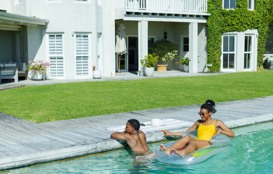 African American couple lounging by pool, man leaning, woman floating on inflatable in swimsuit