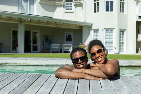 African American couple leaning in swimwear on pool deck smiling with sunglasses by backyard pool