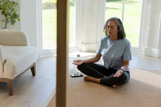 African American woman meditating cross-legged on mat at home in gray tee and leggings with phone