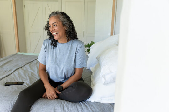 African American senior woman sitting on bed in gray tee and leggings smiling at smartphone