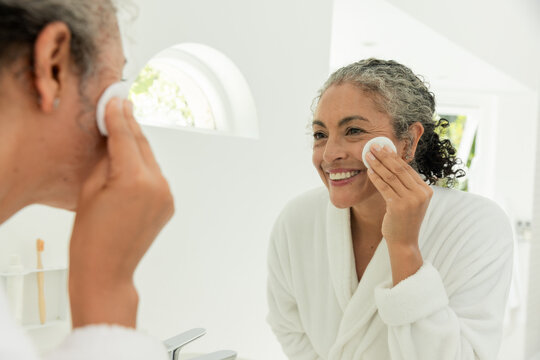 Vertical video: Window light drawing woman in white bathrobe pressing cotton pad to cheek at mirror