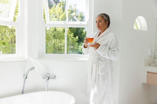 Senior African American woman in white robe standing right in bath holding glass teacup, copy space