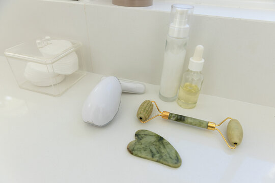 Jade roller and gua sha resting on glossy bathroom countertop with oil dropper, lotion pump, device