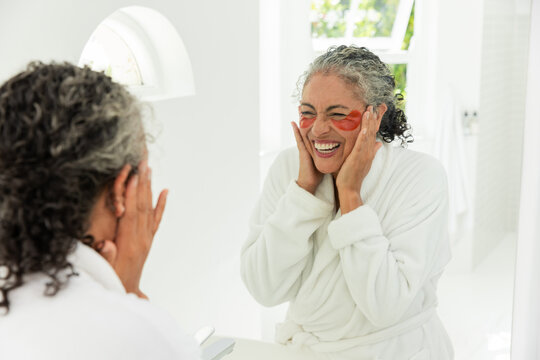 Woman checking mirror after applying gel eye patches, smiling and pressing cheeks for skincare
