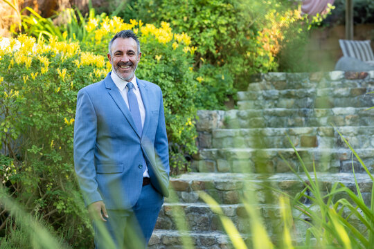 Middle-aged man standing in landscaped garden wearing light blue suit and tie near stone staircase
