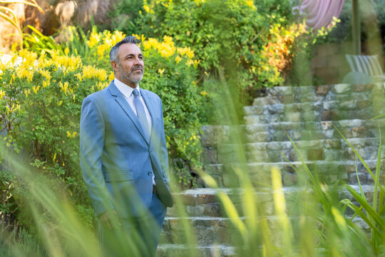 Middle-aged man standing in garden wearing light blue suit, gray tie, pocket square, gazing upward