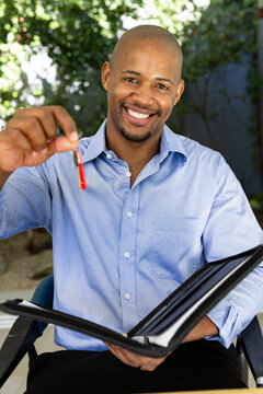 Adult African man smiling, wearing blue shirt and black pants, holding red key, portfolio on patio