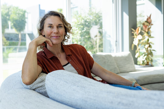 Mid-adult woman reclining on gray sectional sofa in living room, wearing rust shirt and smartwatch