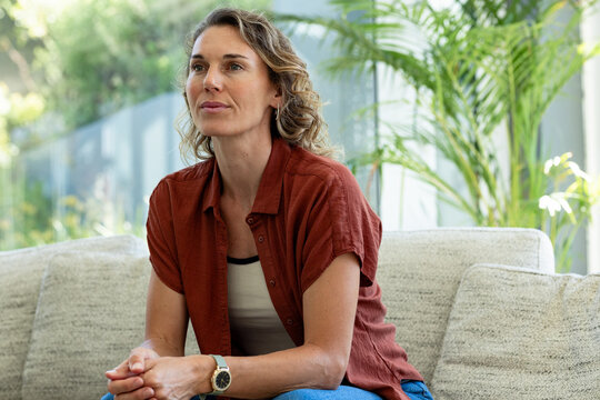 Mid-adult woman sitting on sofa in sunroom wearing rust shirt watch gazing through glass at garden