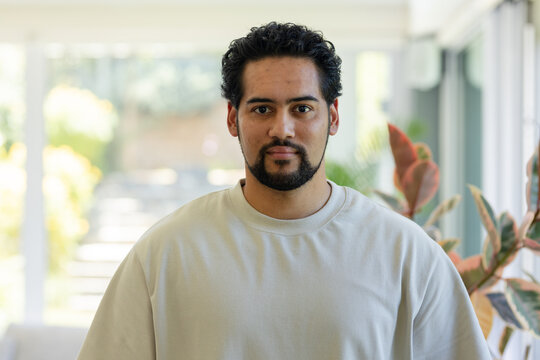 African American man posing in sunroom in beige crewneck, plants, glass doors showing blurred steps