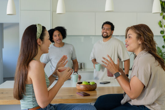 Friends sitting on stools in modern kitchen holding mugs near bowl of green apples and smartwatch