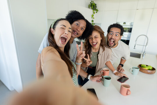 Diverse friends making playful faces for selfie in modern kitchen with smartphones, pastel mugs