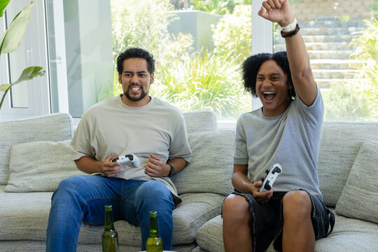 African American male pals sitting on light gray couch, gaming with white gamepads, green bottles