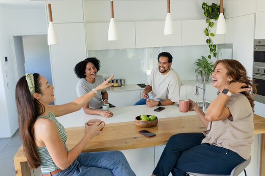 Diverse friends laughing and chatting while holding ceramic mugs around wooden kitchen island