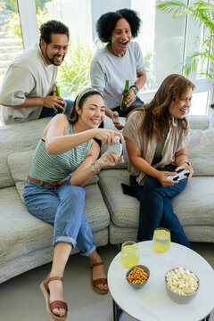 Diverse friends sitting at home on sectional couch, playing game, holding controllers and beers