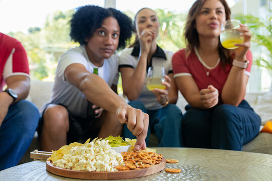Diverse friends reaching across couch at home picking pretzel from board with guacamole, drinks