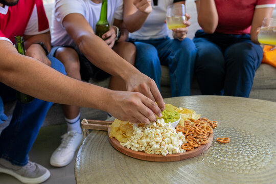 Friends sitting on sofa at home reaching for snacks on serving board with popcorn and dip