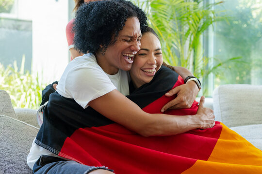 Vertical video: Laughing woman on left hugging friend on couch at home, sharing warmth, red blanket