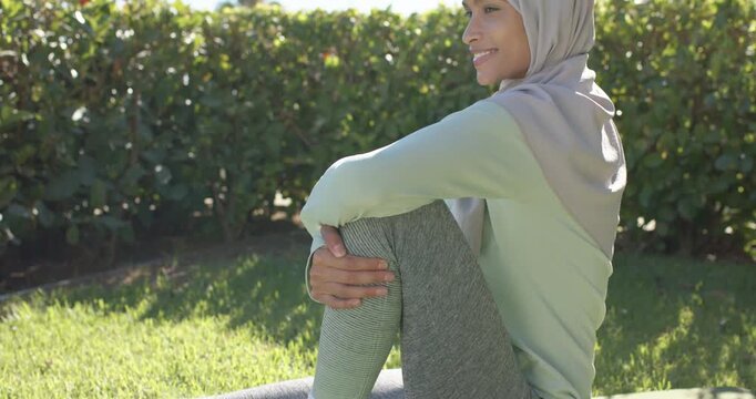 Adult hijab-wearing woman pulling bent knee on green mat for cooldown on lawn as camera panning