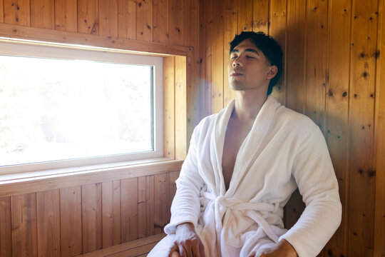Asian man reclining on wooden bench in wood-paneled corner near sunlit window, wearing white robe
