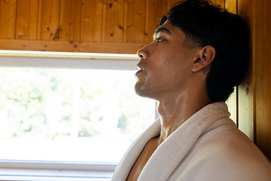 Asian man leaning in sauna against wood-paneling by large window sweating in white robe, copy space