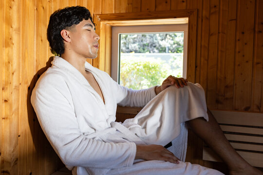 Asian man leaning on wooden bench in sauna wearing white bathrobe holding phone by window