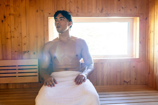 Asian man sitting on wooden bench in sauna, wearing white towel and sweating under backlit window