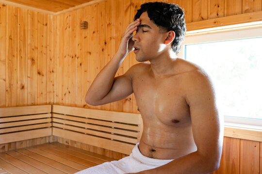 Asian adult male sitting in sauna room holding forehead, resting on wooden bench with white towel