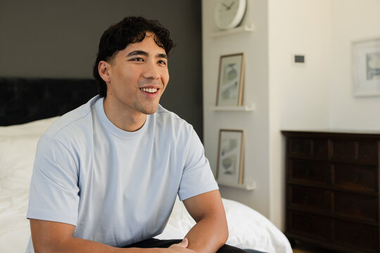 Asian man sitting on edge of bed in bedroom, wearing light blue T-shirt, admiring botanical prints