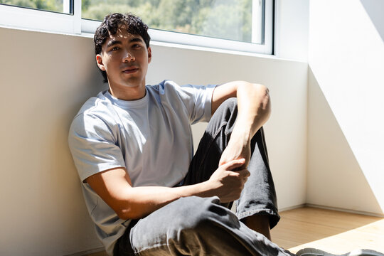 Man in his twenties sitting on hardwood floor leaning under horizontal window with sunlight patch