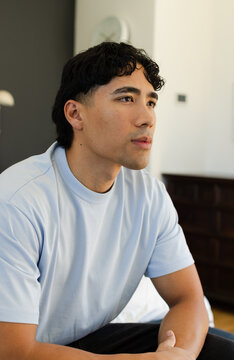 Asian African American man wearing light blue T-shirt, sitting on bed edge, looking at clock