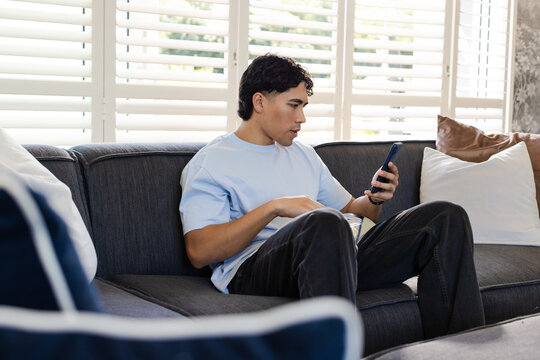Asian adult male sitting on gray sofa at home wearing t-shirt holding smartphone and snack bowl