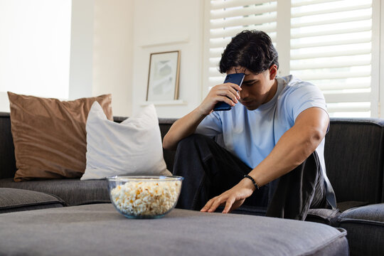 Adult Asian man sitting on gray sofa at home, holding smartphone to forehead, reaching popcorn bowl