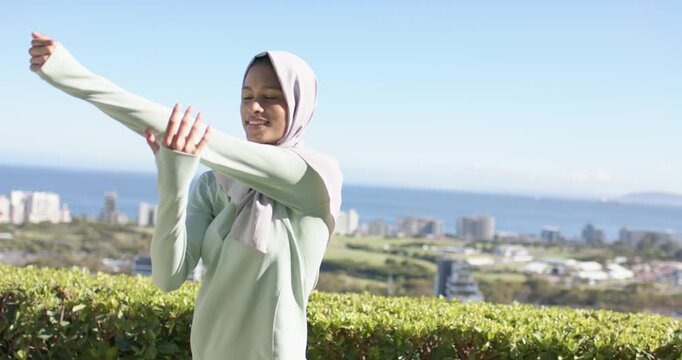 Woman wearing hijab and mint top doing shoulder stretches at coastal view near hedge before walk