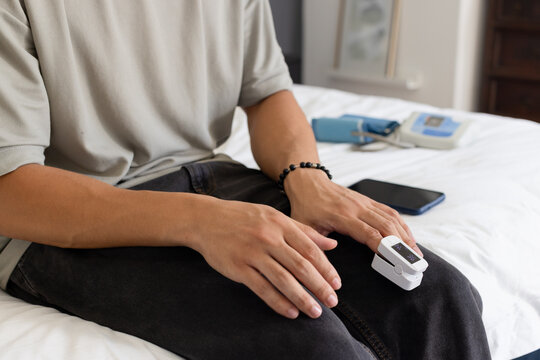 Asian man wearing light gray T-shirt sitting on bed monitoring oxygen with pulse oximeter