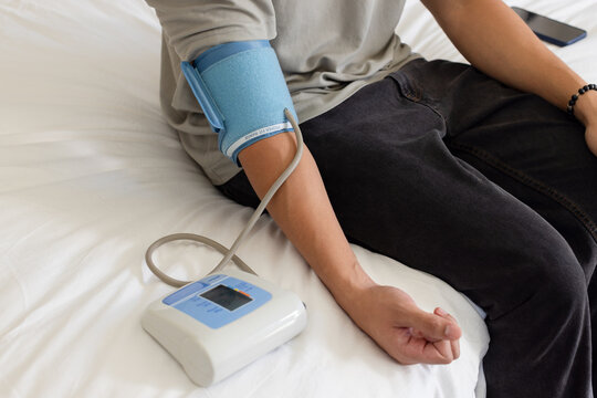 Asian man wearing gray shirt sitting on bed measuring blood-pressure with cuff, monitor, phone
