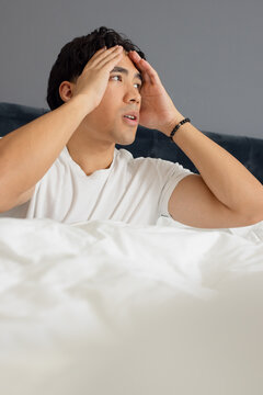 Adult Asian man sitting on bed holding temples, wearing white tee beads, dark headboard gray wall