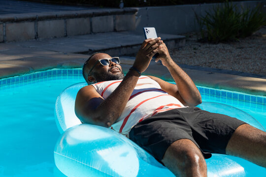 Mid-adult African man reclining on pool lounger at home pool in striped tank holding smartphone