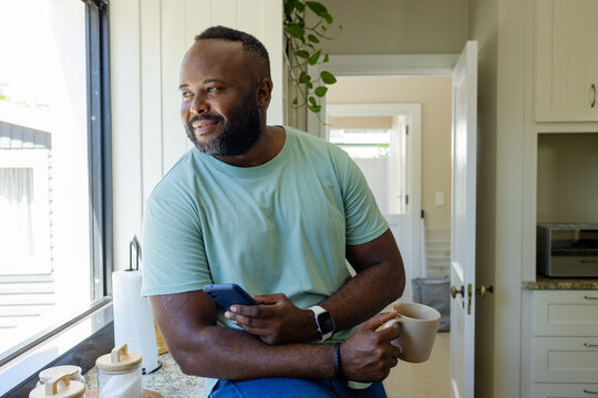 Mid adult African American man sitting on kitchen countertop near window holding smartphone and mug