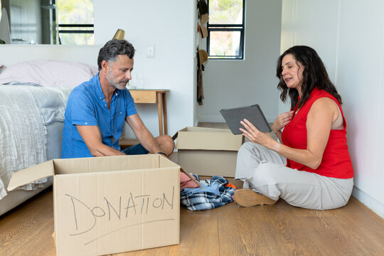 Donation box is overflowing with clothing on hardwood floor near bed, tablet visible